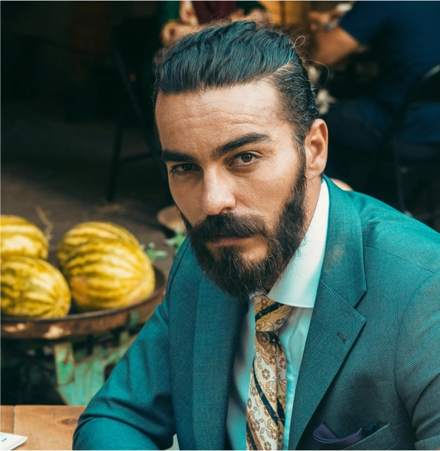 Confident man with a well-groomed beard and slick hairstyle in a teal suit, symbolizing the refined art of modern barbering and timeless masculine elegance.